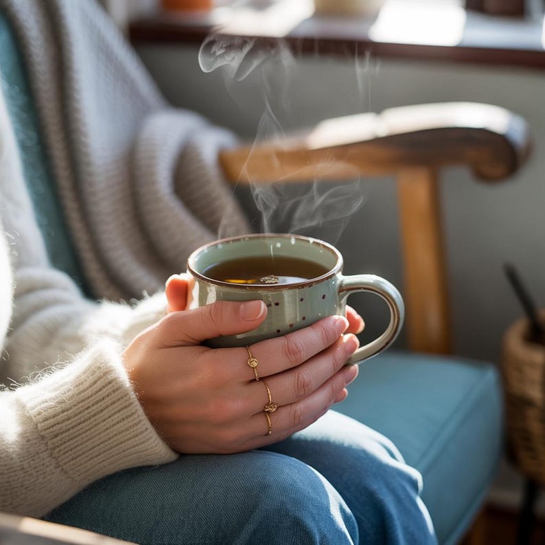 May include: A light blue mug with brown polka dots and a brown rim, filled with steaming tea. The mug is held in both hands. The person is wearing a cream sweater and blue jeans. Steam rises from the tea, suggesting warmth and comfort.