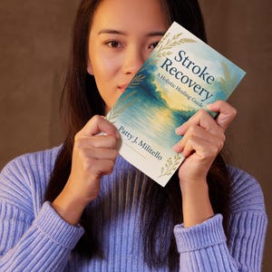 Può includere: Una persona tiene in mano un libro intitolato "Stroke Recovery: A Holistic Healing Guide" davanti al viso. La copertina del libro presenta un paesaggio ad acquerello blu e oro. Il nome dell'autore, Patty J. Militello, è visibile.