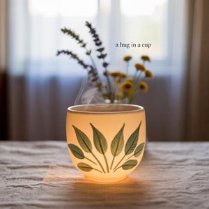 May include: A lit white ceramic cup with a leaf design, filled with a warm drink and steam rising. The text "a hug in a cup" is above the cup. A vase of flowers is in the background.