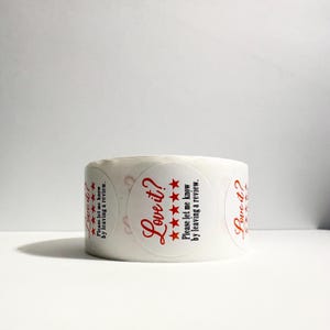 May include: A roll of circular white adhesive stickers featuring red text that reads Love it? followed by five red stars and the text Please let me know by leaving a review. The stickers are arranged in a continuous line on a white backing roll.