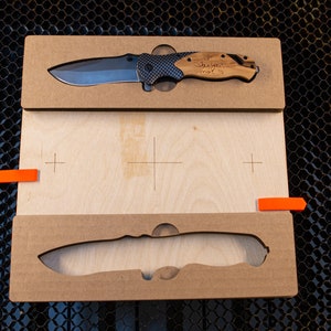 May include: A wooden box with a cutout for a knife. The knife has a black and grey handle with a wood inlay. The knife is engraved with the text "Steve your mom".