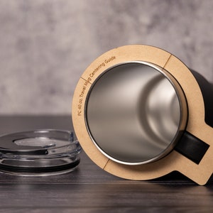May include: A black travel mug with a brown centering guide that says "PC 40 oz. Travel Mug Centering Guide". The mug is open and the lid is next to it. The lid is clear plastic with a black rubber seal.