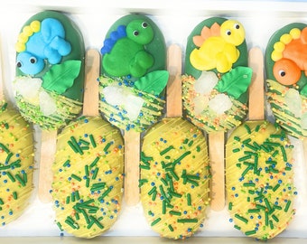 May include: A box of dinosaur-themed cake pops. The pops feature green, yellow, blue, and orange dinosaur decorations on a green background. The pops are on wooden sticks and decorated with sprinkles.