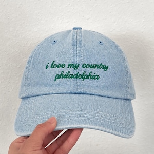 May include: A light blue denim baseball cap with a curved brim and a button on top. The front of the cap features embroidered dark green text that reads "i love my country philadelphia" in a lowercase, cursive-style font across two lines.