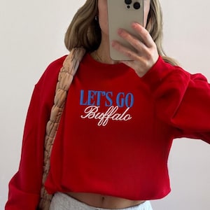 May include: Red sweatshirt with the text "Let's Go Buffalo" in blue and white. The person is wearing gray sweatpants, white sneakers, and a beige cap. A beige bag is over the shoulder.