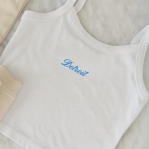 Op de afbeelding: Witte tanktop met dunne bandjes en het woord "Detroit" in blauw geborduurd. De top is gemaakt van een zachte, lichte stof. Andere witte kledingstukken zijn zichtbaar op de achtergrond.