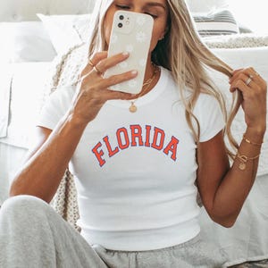 May include: White t-shirt with the word "FLORIDA" in red arched lettering. The person is wearing grey sweatpants and white fluffy slippers. The person is holding a white phone with a floral design.