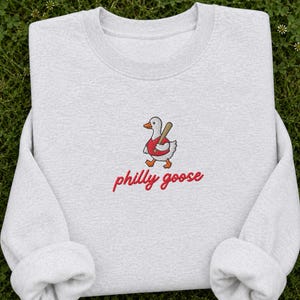 Può includere: Una felpa girocollo grigio chiaro con un ricamo raffigurante un'oca bianca che indossa un gilet rosso e trasporta una mazza da baseball marrone. Sotto l'oca, il testo philly goose è ricamato in corsivo rosso.