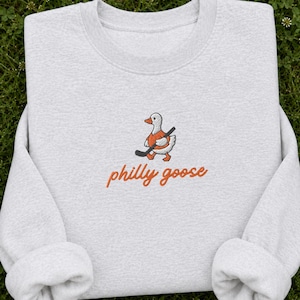 Könnte beinhalten: Ein hellgraues Sweatshirt mit Rundhalsausschnitt und einer Stickerei, die eine weiße Gans in einem orangefarbenen Trikot mit einem schwarzen Hockeyschläger zeigt, sowie dem orangefarbenen Schriftzug "philly goose" darunter.