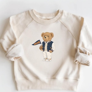 May include: A cream-colored long-sleeve sweatshirt featuring a central graphic of a brown teddy bear wearing a navy blue and white varsity jacket, white pants, and white shoes, while holding a navy blue pennant with the word CHICAGO in orange.