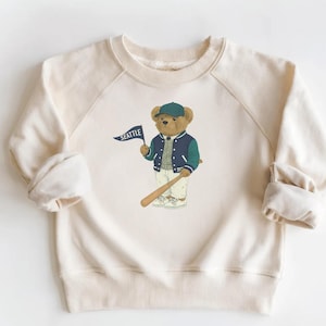 May include: Cream-colored sweatshirt with a baseball-themed bear graphic. The bear wears a blue and green jacket and a cap, holding a baseball bat and a flag that says "SEATTLE". The sweatshirt has raglan sleeves.