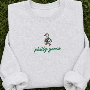 May include: A light gray crewneck sweatshirt featuring an embroidered graphic of a white goose wearing a green jersey and holding a brown football. Below the goose, the text "philly goose" is embroidered in a green cursive script font.
Philadelphia Football, Embroidered Crewneck Sweatshirt, T-Shirt Embroidery, Philly Goose, Silly Goose
philly goose philly goose sweatshirt philadelphia philly goose shirt retro philadelphia designs philly crewneck