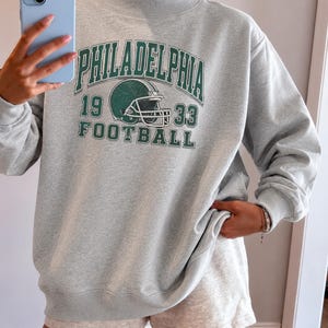 May include: A light grey mock neck sweatshirt featuring a dark green graphic print that reads PHILADELPHIA FOOTBALL 1933 with an illustration of a football helmet. The long-sleeved pullover has ribbed cuffs and a ribbed hem, constructed from a soft fabric.