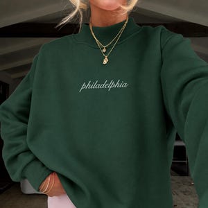 May include: A dark forest green long-sleeve sweatshirt featuring a mock neck collar and the word philadelphia embroidered in white cursive script across the center chest, paired with light pastel pink shorts.
