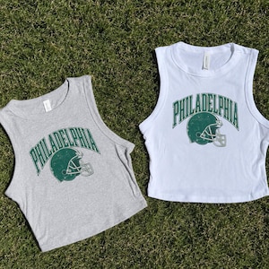 Może przedstawiać: Dwa bezrękawniki typu crop top, jeden w kolorze szarego melanżu, a drugi biały, każdy z ciemnozieloną grafiką kasku do futbolu amerykańskiego i napisem PHILADELPHIA łukowatą czcionką. Wzory mają postarzany, przetarty wygląd.