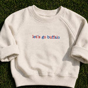 May include: A cream-colored crewneck sweatshirt with long sleeves and ribbed cuffs. The front features the text 'let's go buffalo' embroidered in red and blue lowercase letters. The garment has a simple, classic design with a round neckline.