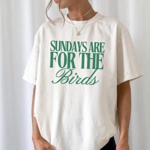 May include: An ivory-coloured t-shirt with the green text "SUNDAYS ARE FOR THE Birds". The shirt has a loose fit and short sleeves. The text "IVORY SIZE XL" is at the bottom right.