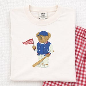 Op de afbeelding: Crèmekleurig T-shirt met een cartoonbeer met een blauwe baseballpet en jas, met een honkbalknuppel en een Chicago-vlag. Het shirt heeft een Comfort Colors-label. Een rood-witte geruite pyjamabroek is zichtbaar.