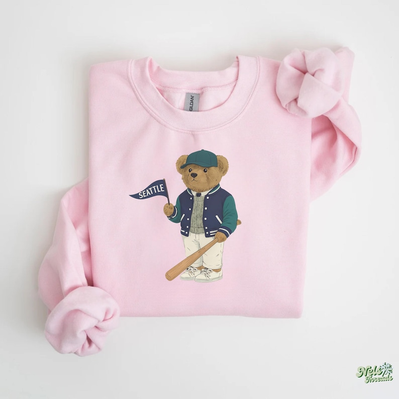 May include: A pale pink sweatshirt with a cartoon bear wearing a baseball cap and jacket, holding a baseball bat and a flag that says "SEATTLE". The bear is wearing a navy blue and teal jacket, white trousers, and trainers.