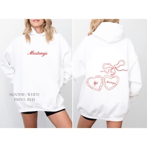 Puede incluir: Sudadera blanca con texto rojo "Mustangs" en la parte delantera. La parte trasera presenta un dise&ntilde;o de medall&oacute;n en forma de coraz&oacute;n rojo con "Go Mustangs". La sudadera con capucha tiene una capucha y un bolsillo delantero.