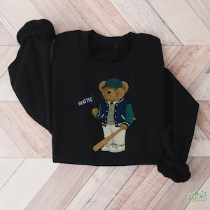 May include: Black sweatshirt with an embroidered bear wearing a baseball cap and jacket, holding a bat and a flag that reads "SEATTLE". The bear is in a baseball uniform. The sweatshirt is made of a soft material.