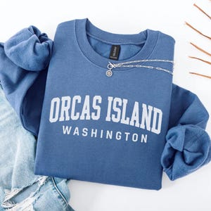 May include: A blue sweatshirt displaying the text "ORCAS ISLAND WASHINGTON" in white. A silver necklace with a pendant is placed on the front. The sweatshirt is folded with a pair of blue jeans.