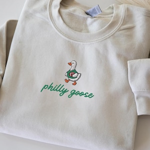 May include: A sand-colored crewneck sweatshirt featuring an embroidered graphic of a white goose wearing a green jersey and holding a brown football. Below the goose, the text "philly goose" is embroidered in green cursive script lettering.
Philadelphia Football, Embroidered Crewneck Sweatshirt, T-Shirt Embroidery, Philly Goose, Silly Goose
philly goose philly goose sweatshirt philadelphia philly goose shirt retro philadelphia designs philly crewneck