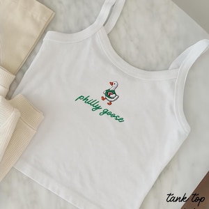 May include: A white tank top featuring an embroidered graphic of a goose wearing a green jersey and holding a football, with the text philly goose in green cursive script below the goose. The garment has thin shoulder straps and a scoop neckline.
Philadelphia Football, Embroidered Crewneck Sweatshirt, T-Shirt Embroidery, Philly Goose, Silly Goose
philly goose philly goose sweatshirt philadelphia philly goose shirt retro philadelphia designs philly crewneck tank top spaghetti straps white tank