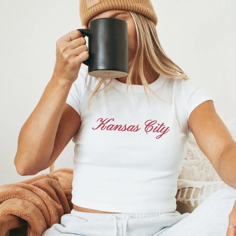 May include: A white crop top with the cursive text "Kansas City" in red. The person is wearing a brown beanie and holding a black mug. The person is sitting in a chair with a brown blanket.