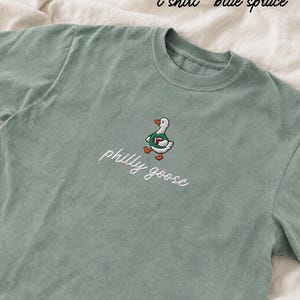May include: A blue spruce t-shirt featuring an embroidered graphic of a white goose wearing a green jersey and holding a brown football. Below the goose, the text philly goose is embroidered in white cursive script. The shirt has a crew neckline.
Philadelphia Football, Embroidered Crewneck Sweatshirt, T-Shirt Embroidery, Philly Goose, Silly Goose
philly goose philly goose sweatshirt philadelphia philly goose shirt retro philadelphia designs philly crewneck