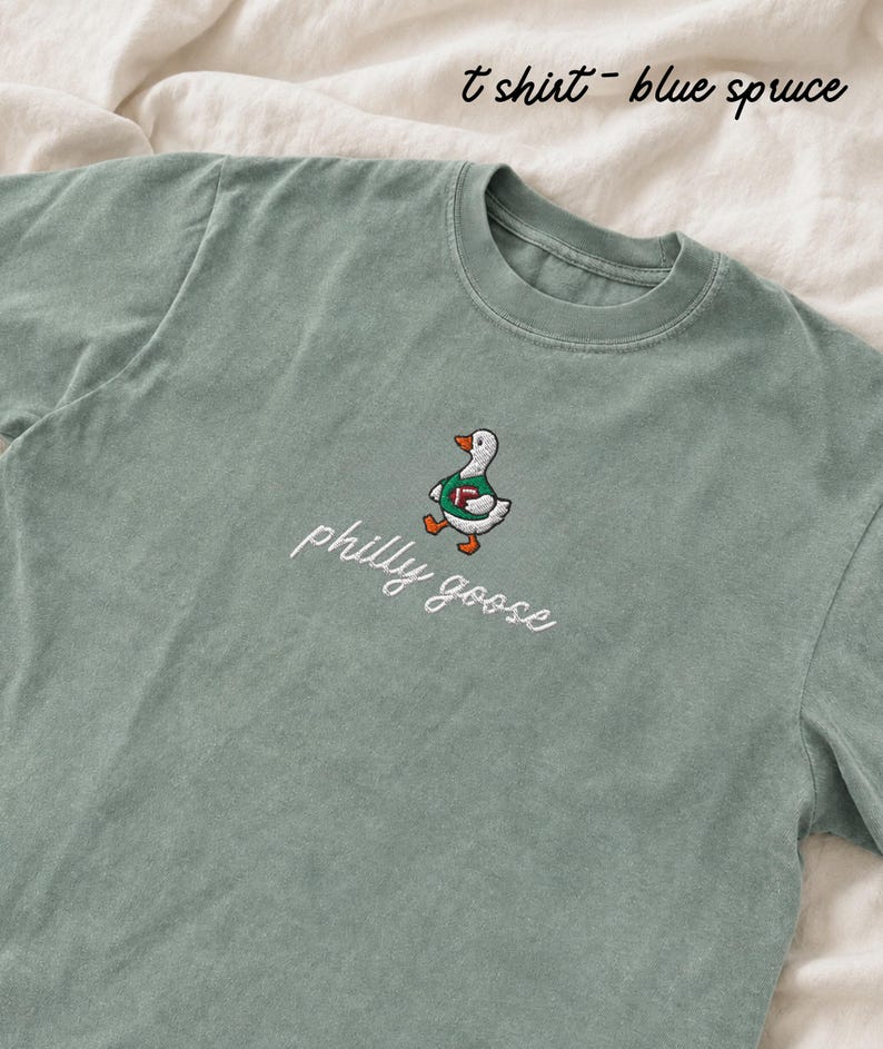 May include: A blue spruce t-shirt featuring an embroidered graphic of a white goose wearing a green jersey and holding a brown football. Below the goose, the text philly goose is embroidered in white cursive script. The shirt has a crew neckline.
Philadelphia Football, Embroidered Crewneck Sweatshirt, T-Shirt Embroidery, Philly Goose, Silly Goose
philly goose philly goose sweatshirt philadelphia philly goose shirt retro philadelphia designs philly crewneck