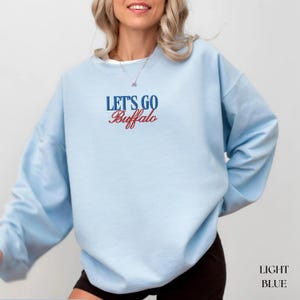 May include: Light blue sweatshirt with the text "LET'S GO Buffalo" in blue and red. The sweatshirt is a solid light blue color. The words "LIGHT BLUE" are printed at the bottom right corner of the image.