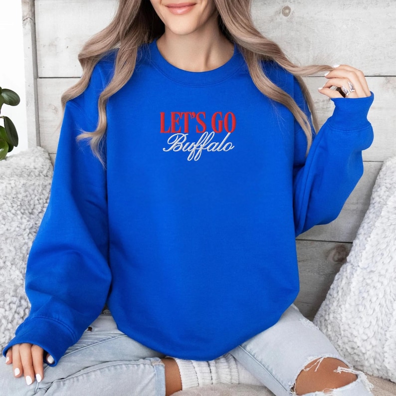 May include: Royal blue sweatshirt with the text "LET'S GO Buffalo" in red and white script. The sweatshirt has a crew neck and long sleeves. The text is centered on the chest.