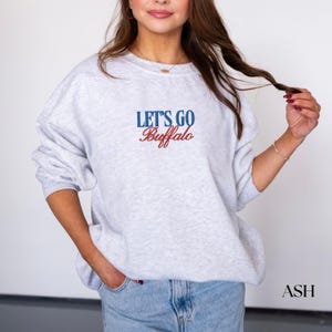 May include: A light gray sweatshirt with the text "LET'S GO Buffalo" in blue and red. The sweatshirt is a crew neck style and has long sleeves. The model is wearing blue jeans.