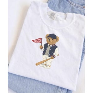 May include: White t-shirt featuring a cartoon bear wearing a baseball cap and jacket, holding a bat and a flag that says "BOSTON". The bear is in a baseball uniform. The shirt is folded on top of a pair of blue jeans.