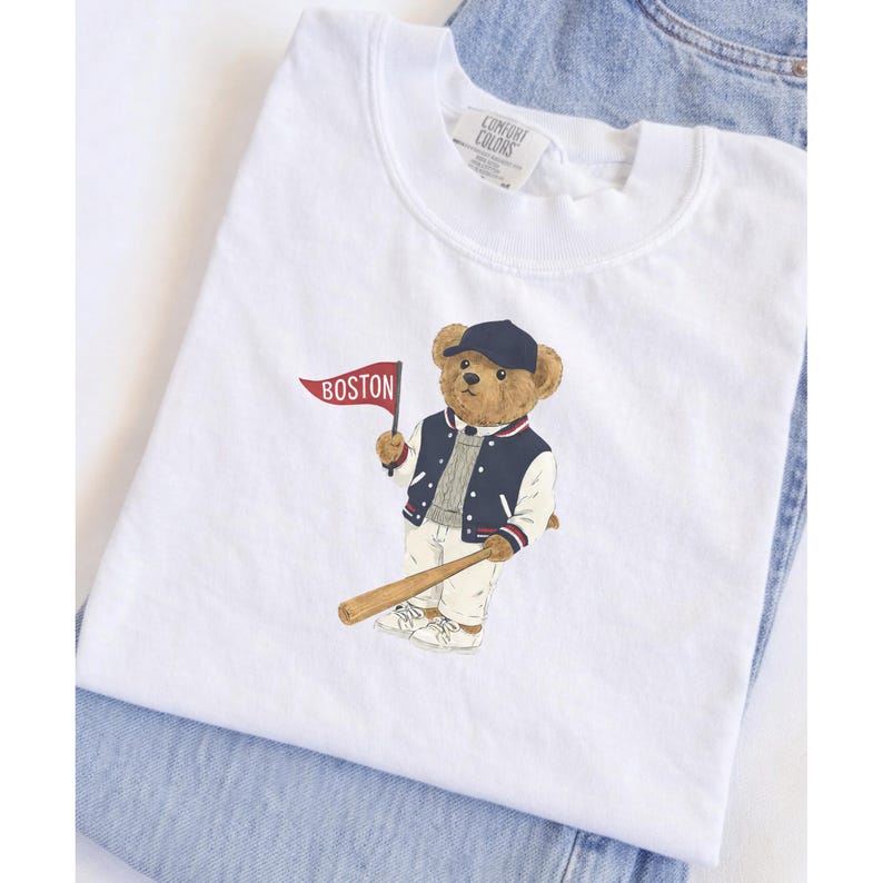 May include: White t-shirt featuring a cartoon bear wearing a baseball cap and jacket, holding a bat and a flag that says "BOSTON". The bear is in a baseball uniform. The shirt is folded on top of a pair of blue jeans.
