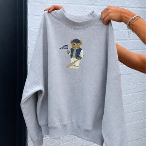 Op de afbeelding: Een lichtgrijs sweatshirt met ronde hals en een centrale afbeelding van een bruine teddybeer met een marineblauwe honkbalpet, een marineblauw-wit varsity-jack en een witte broek, die een marineblauwe wimpel vasthoudt met de tekst NEW YORK.