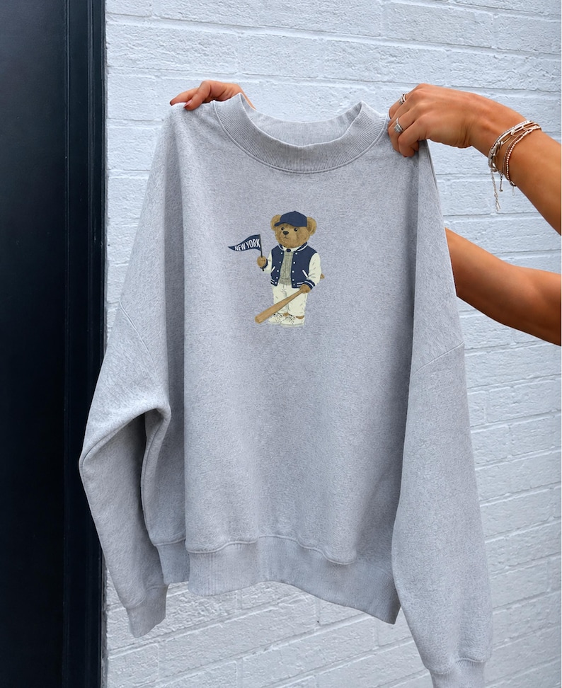 Op de afbeelding: Een lichtgrijs sweatshirt met ronde hals en een centrale afbeelding van een bruine teddybeer met een marineblauwe honkbalpet, een marineblauw-wit varsity-jack en een witte broek, die een marineblauwe wimpel vasthoudt met de tekst NEW YORK.