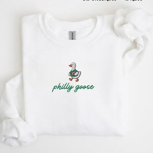 May include: A white crewneck sweatshirt featuring an embroidered graphic of a goose wearing a green jersey and holding a football. Below the goose, the text philly goose is embroidered in green script. The text sweatshirt - white appears at the top.
Philadelphia Football, Embroidered Crewneck Sweatshirt, T-Shirt Embroidery, Philly Goose, Silly Goose
philly goose philly goose sweatshirt philadelphia philly goose shirt retro philadelphia designs philly crewneck