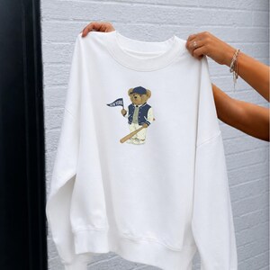 Op de afbeelding: Een wit sweatshirt met ronde hals voorzien van een afbeelding van een teddybeer met een marineblauwe baseballpet, een marineblauw-wit varsity-jack en een witte broek. De beer houdt een klein marineblauw wimpeltje vast met de tekst NEW YORK.
