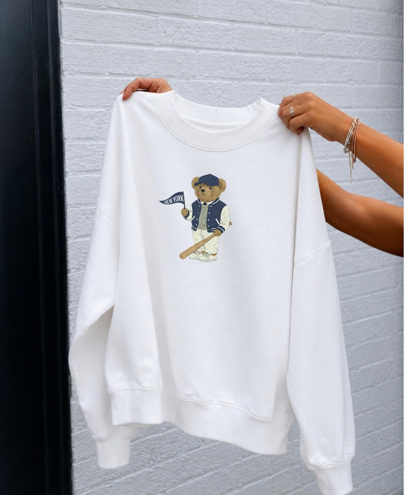 Op de afbeelding: Een wit sweatshirt met ronde hals voorzien van een afbeelding van een teddybeer met een marineblauwe baseballpet, een marineblauw-wit varsity-jack en een witte broek. De beer houdt een klein marineblauw wimpeltje vast met de tekst NEW YORK.