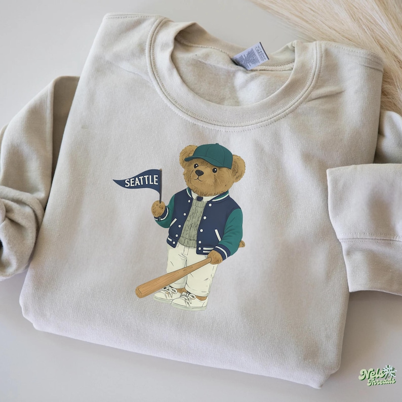 May include: A cream-coloured sweatshirt with a cartoon bear wearing a baseball cap and jacket, holding a baseball bat and a flag that says "SEATTLE". The bear has a green and navy blue jacket and cap. The sweatshirt is a neutral colour.