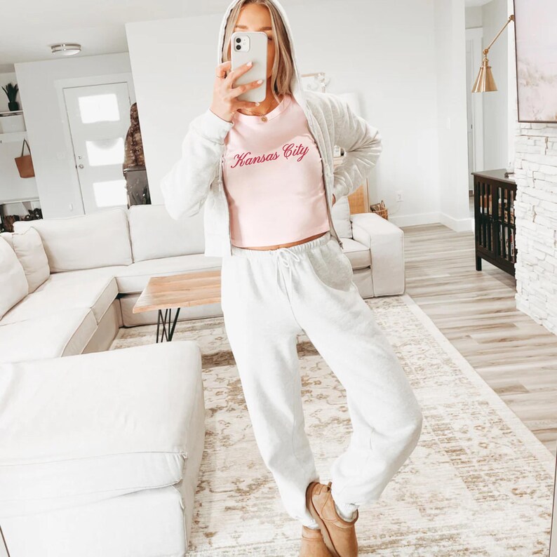 May include: A light pink crop top with the text "Kansas City" in a cursive font. The top is paired with light gray sweatpants and a matching zip-up hoodie. The outfit is completed with brown boots.