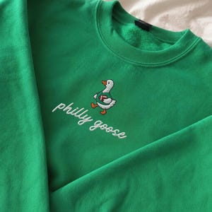 May include: An Irish green sweatshirt featuring an embroidered graphic of a white goose wearing a green jersey and holding a brown football. Below the goose, the text philly goose is embroidered in white cursive script. The fabric appears soft.
Philadelphia Football, Embroidered Crewneck Sweatshirt, T-Shirt Embroidery, Philly Goose, Silly Goose
philly goose philly goose sweatshirt philadelphia philly goose shirt retro philadelphia designs philly crewneck