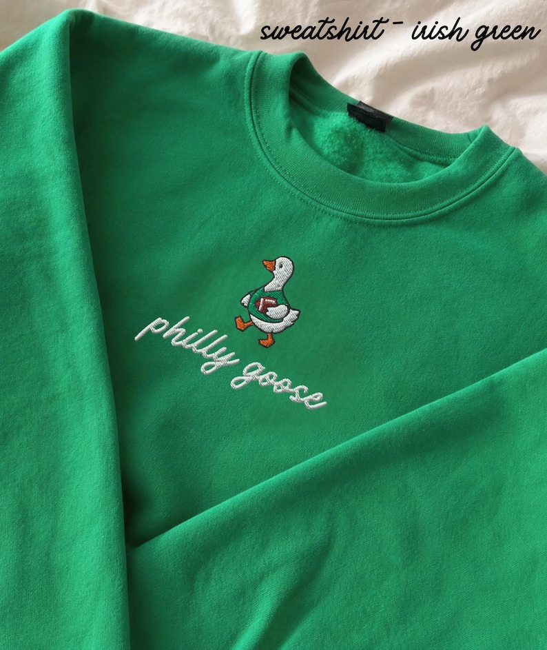 May include: An Irish green sweatshirt featuring an embroidered graphic of a white goose wearing a green jersey and holding a brown football. Below the goose, the text philly goose is embroidered in white cursive script. The fabric appears soft.
Philadelphia Football, Embroidered Crewneck Sweatshirt, T-Shirt Embroidery, Philly Goose, Silly Goose
philly goose philly goose sweatshirt philadelphia philly goose shirt retro philadelphia designs philly crewneck