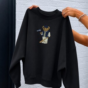 Op de afbeelding: Een zwart sweatshirt met ronde hals en een afbeelding van een teddybeer met een blauwe honkbalpet, een marineblauw-wit varsity-jack en een witte broek. De beer houdt een houten honkbalknuppel en een blauw vlaggetje met de tekst NEW YORK vast.