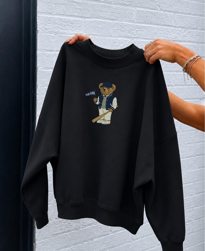 Op de afbeelding: Een zwart sweatshirt met ronde hals en een afbeelding van een teddybeer met een blauwe honkbalpet, een marineblauw-wit varsity-jack en een witte broek. De beer houdt een houten honkbalknuppel en een blauw vlaggetje met de tekst NEW YORK vast.