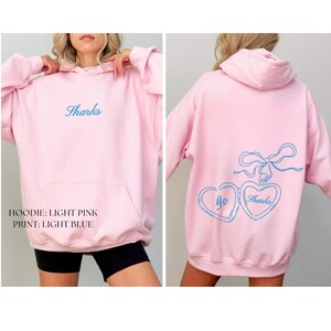 Puede incluir: Sudadera con capucha rosa claro con bolsillo delantero y capucha. La palabra "Sharks" est&aacute; bordada en azul claro en la parte delantera. La parte trasera presenta un dise&ntilde;o azul claro de dos medallones en forma de coraz&oacute;n con las palabras "go" y "Sharks".