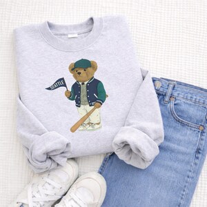 May include: A light grey sweatshirt with a cartoon bear in a baseball jacket and cap, holding a bat and a flag reading "SEATTLE". The sweatshirt is styled with blue jeans and white trainers.