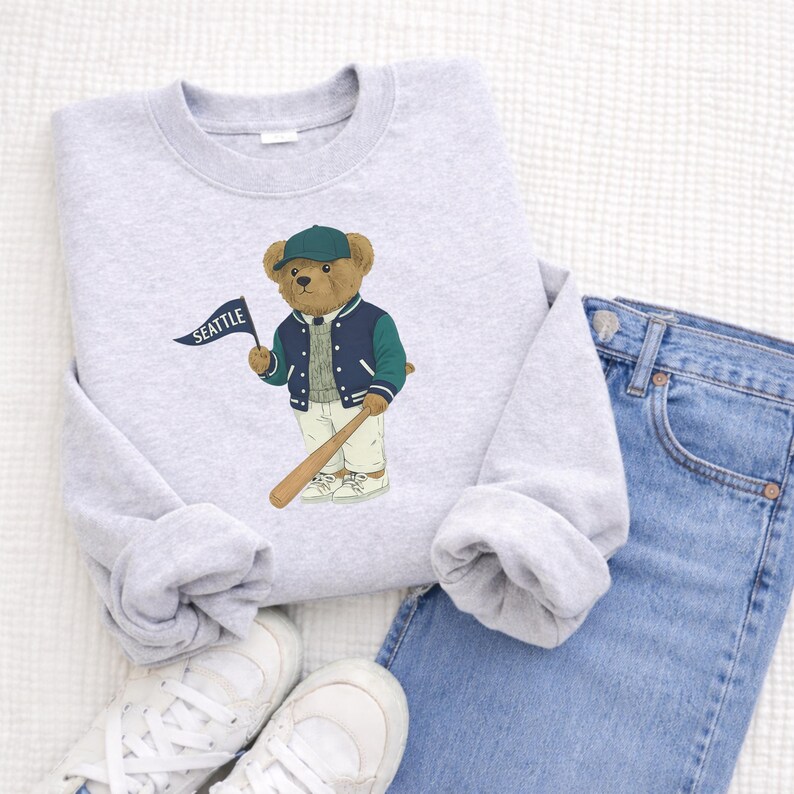 May include: A light grey sweatshirt with a cartoon bear in a baseball jacket and cap, holding a bat and a flag reading "SEATTLE". The sweatshirt is styled with blue jeans and white trainers.